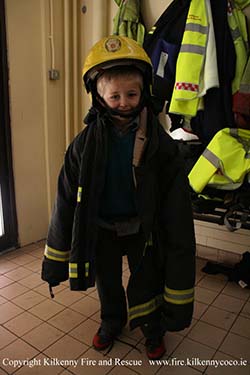 Vaikas bando apsirengti ugniagesių uniformą
